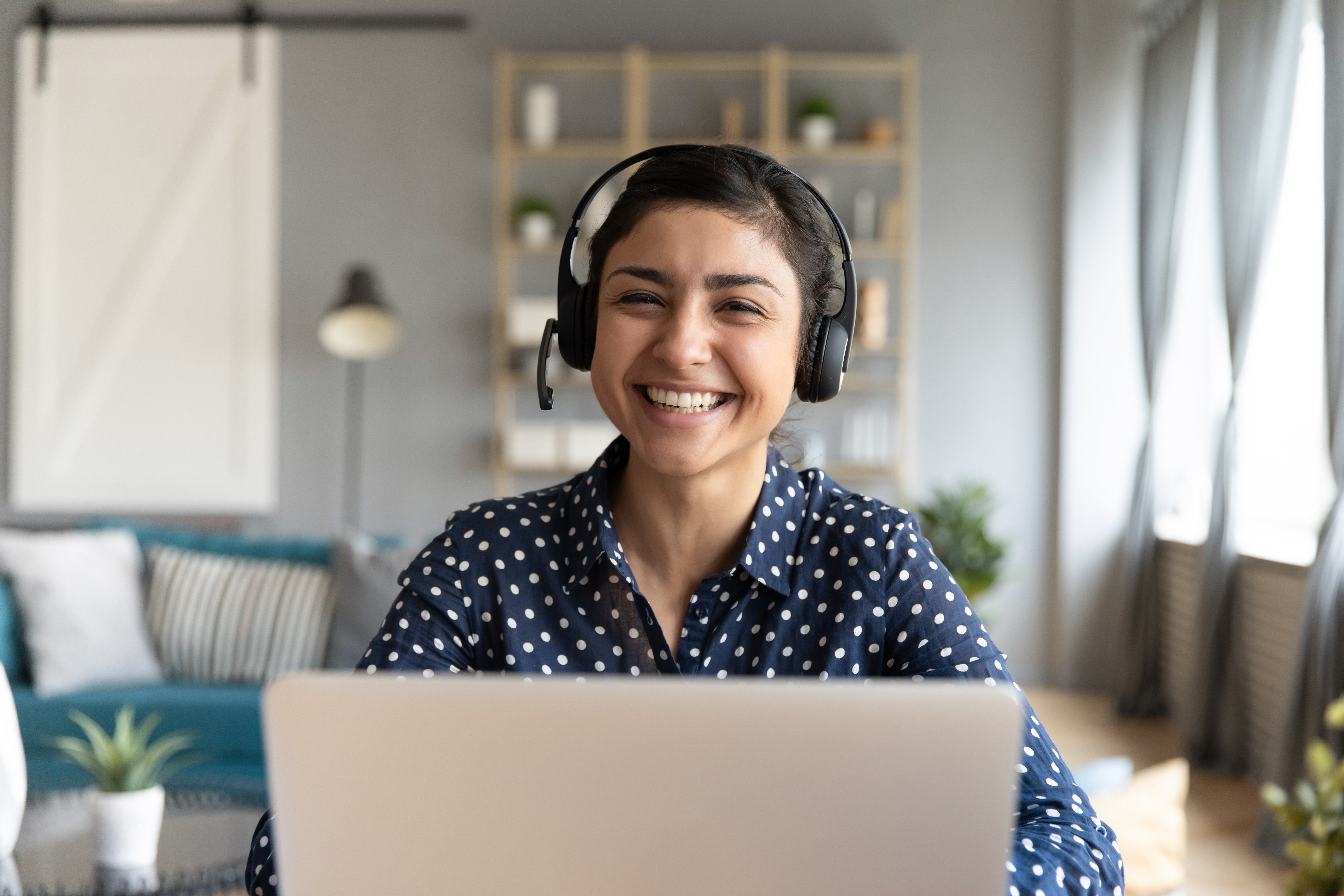 Head shot cheerful smiling pretty hindu ethnic girl sitting at table with computer, wearing headphones with mic, looking at camera. Happy indian woman professional tutor educating client online.