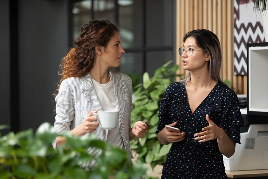 Diverse employees chatting during coffee break, walking in modern office, Asian businesswoman wearing glasses sharing ideas, discussing project with colleague, having pleasant conversation